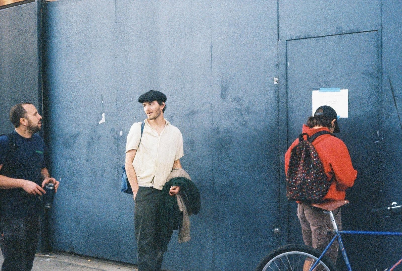Louis in a flat cap against a blue hoarding, a friend beside him and a cyclist at the door