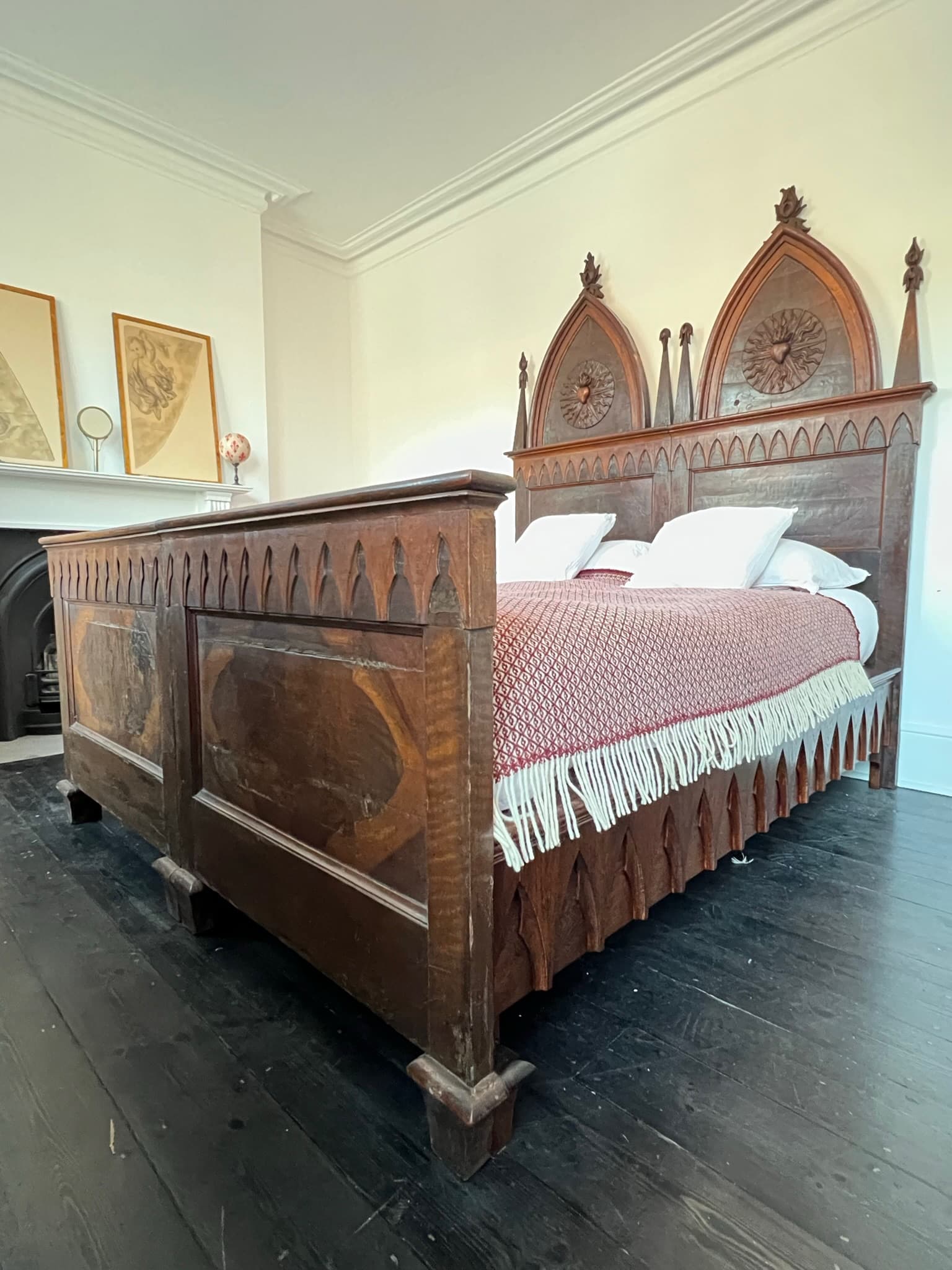 The bed made up in a red wool throw, its carved headboard of two pointed arches beyond the panelled footboard